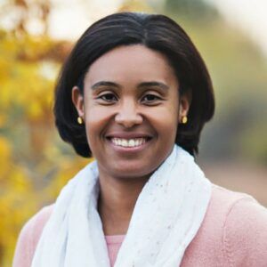 Nichole Tiggs Individual with white scarf and pink sweater smiling. Background is trees.